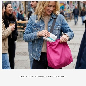 May include: A woman in a denim jacket and tan sweater is putting an item into a bright pink drawstring bag. The bag is slung over her shoulder. The text at the bottom of the image reads "LEICHT GETRAGEN IN DER TASCHE."