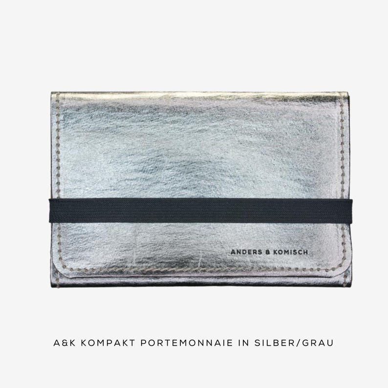 May include: A silver wallet with a black elastic band closure. The wallet has a textured surface and the text "ANDERS & KOMISCH" is printed on the front. The text "A&K KOMPAKT PORTEMONNAIE IN SILBER/GRAU" is printed below the wallet.