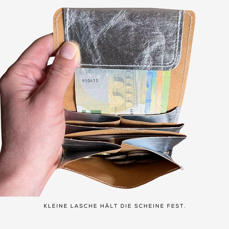 May include: A silver and tan leather wallet, open to display cash and coins. The wallet has multiple compartments and a silver metallic finish on the exterior. The text "KLEINE LASCHE H&Auml;LT DIE SCHEINE FEST" is visible at the bottom.