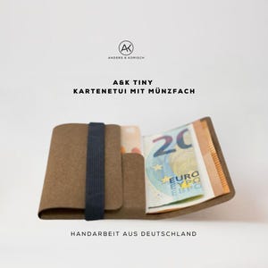 Op de afbeelding: Een bruine leren portemonnee met een zwarte elastische band, met een creditcard en een biljet van 20 euro. De portemonnee heeft de tekst "A&K TINY KARTENETUI MIT MÜNZFACH" en "HANDARBEIT AUS DEUTSCHLAND".