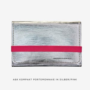 May include: Silver wallet with a bright pink elastic band closure. The wallet has a textured surface and the text "ANDERS & KOMISCH" is printed on the front. The wallet is rectangular and has stitched edges.
