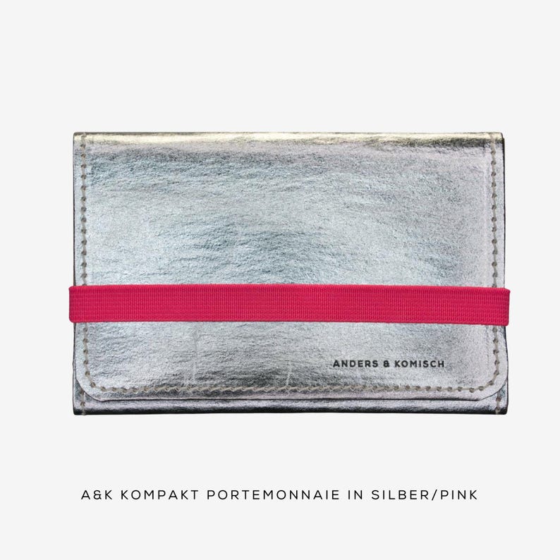 May include: Silver wallet with a bright pink elastic band closure. The wallet has a textured surface and the text "ANDERS & KOMISCH" is printed on the front. The wallet is rectangular and has stitched edges.