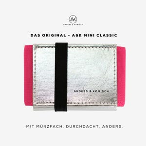 Könnte beinhalten: Ein silbernes Portemonnaie mit schwarzem Gummibandverschluss und leuchtend pinken Seiten. Der Text "A&K MINI CLASSIC" ist sichtbar, zusammen mit dem Markennamen "ANDERS & KOMISCH". Das Portemonnaie hat ein minimalistisches Design.