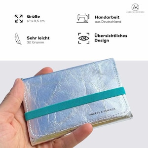 May include: Silver wallet with a teal elastic band, measuring 12 x 8.5 cm. The wallet is handmade in Germany and weighs 32 grams. The text "ANDERS & KOMISCH" is printed on the wallet.