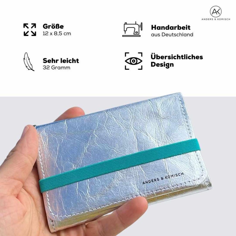 May include: Silver wallet with a teal elastic band, measuring 12 x 8.5 cm. The wallet is handmade in Germany and weighs 32 grams. The text "ANDERS & KOMISCH" is printed on the wallet.