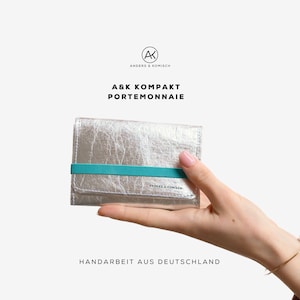 Può includere: Un portafoglio argentato con un elastico turchese è tenuto in mano. Il portafoglio ha un aspetto strutturato e il testo "A&K KOMPAKT PORTEMONNAIE" è visibile. Sono visibili anche il logo del marchio e "HANDARBEIT AUS DEUTSCHLAND".