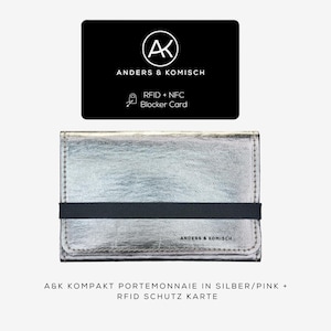 May include: A silver wallet with a black elastic band and a black card with the text "ANDERS & KOMISCH" and "RFID + NFC Blocker Card". The wallet has the text "ANDERS & KOMISCH" on it. The wallet is a compact size.