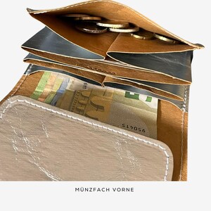 May include: A bi-fold wallet with multiple compartments. The wallet is open, revealing a section with coins and another with folded bills. The exterior is a light tan color, with a contrasting dark gray interior. The text "M&Uuml;NZFACH VORNE" is visible at the bottom.