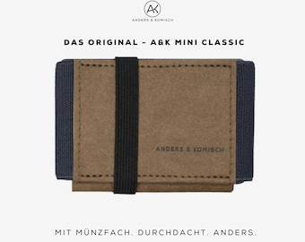 Puede incluir: Una cartera marrón con una banda elástica negra y paneles laterales azul oscuro. El texto "ANDERS & KOMISCH" está impreso en el frente. Arriba, el texto dice "DAS ORIGINAL - A&K MINI CLASSIC" y debajo "MIT MÜNZFACH. DURCHDACHT. ANDERS."