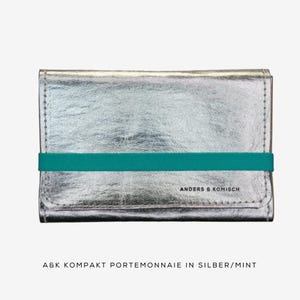 May include: A silver wallet with a teal elastic band. The wallet has a textured metallic finish and is rectangular in shape. The text "ANDERS & KOMISCH" is printed on the front.