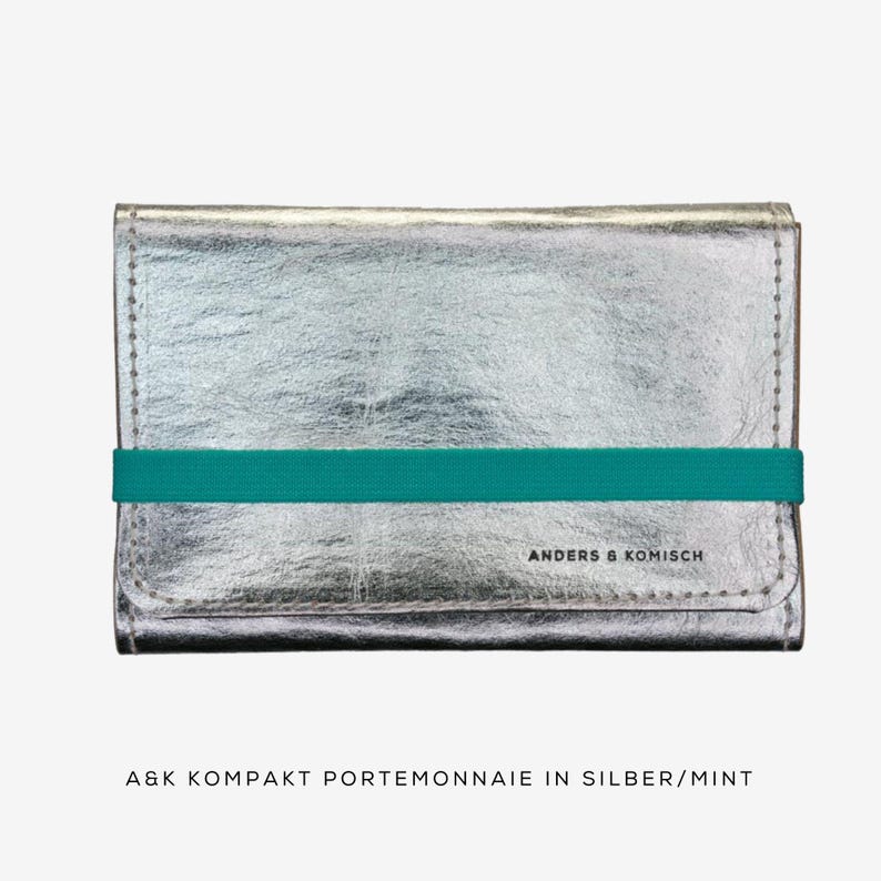 May include: A silver wallet with a teal elastic band. The wallet has a textured metallic finish and is rectangular in shape. The text "ANDERS & KOMISCH" is printed on the front.