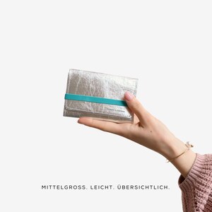May include: A silver wallet with a textured finish and a turquoise elastic band. The wallet is held in a hand against a white background. The text "MITTELGROSS. LEICHT. &Uuml;BERSICHTLICH." is at the bottom.
