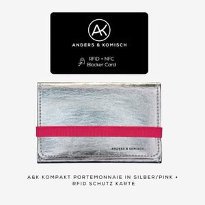 May include: A silver wallet with a bright pink elastic band, and a black card with the text "ANDERS & KOMISCH" and "RFID + NFC Blocker Card". The wallet has the text "ANDERS & KOMISCH" printed on it.