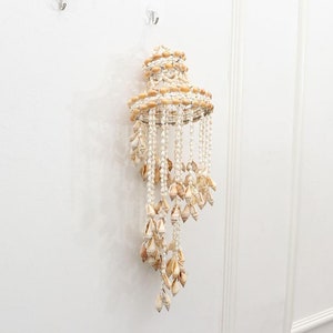 May include: A decorative seashell chandelier with a tiered design. The chandelier is made of various seashells in shades of cream and tan, strung together to create a cascading effect. It hangs from a white wall.