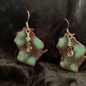 Op de afbeelding: Een paar oorbellen in de vorm van de staat Wisconsin, met een groene en bruine patina. Elke oorbel heeft een klein groen kristal en een gouden draadspiraal.