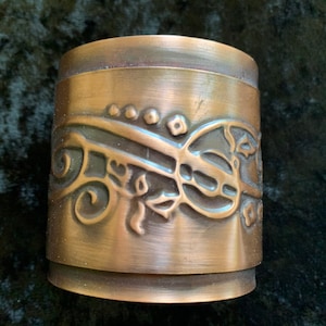 May include: A copper-coloured metal cup with an embossed design of swirling vines and flowers. The cup has a smooth, polished finish.