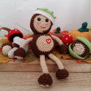 May include: A crocheted doll with a brown body, beige legs and arms, and a green hat. The doll is sitting on a wooden surface with crocheted mushrooms, pumpkins, and acorns.