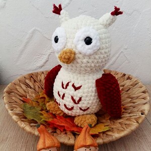 Puede incluir: Un búho de crochet blanco con detalles rojos y naranjas se sienta en una cesta tejida con hojas de otoño. El búho tiene ojos grandes y negros y un pequeño pico naranja.