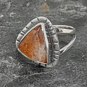 Puede incluir: Un anillo de plata con una piedra triangular de color marrón anaranjado. La piedra está engastada en un bisel plateado texturizado. La banda del anillo es de plata pulida lisa. Probablemente sea una joya.