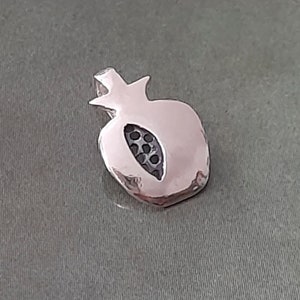 Puede incluir: Un amuleto de plata con forma de granada. El amuleto tiene un diseño detallado con un centro recortado.