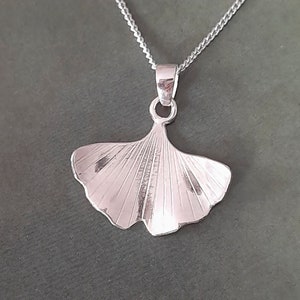 Puede incluir: Collar con colgante plateado con diseño de hoja de ginkgo. El colgante, con líneas grabadas, cuelga de una delicada cadena. El collar se muestra sobre un fondo verde apagado. La cadena y el colgante son plateados.