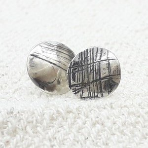 Puede incluir: Un par de pendientes de plata con un diseño texturizado y abstracto. Los pendientes son redondos y tienen un acabado ligeramente martillado.