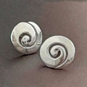 Puede incluir: Un par de pendientes de botón plateados. Cada pendiente presenta un diseño en espiral dentro de una forma circular. Los pendientes están sobre un fondo marrón oscuro, resaltando su brillo metálico y sus intrincados detalles. Joyas.