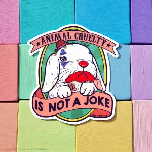 May include: A white rabbit wearing a red nose and a hat with a star on it. The rabbit is holding a red flower and is looking at the camera. The text "Animal Cruelty is not a joke" is written on a banner around the rabbit. The background is a light blue color.