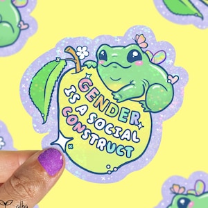 May include: A sticker featuring a green frog with butterfly wings perched on a pear. The pear has the text "GENDER IS A SOCIAL CONSTRUCT" in pastel colors. The sticker has a glittery border and is on a yellow background.