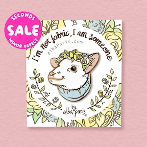 May include: A white enamel pin with a cartoon opossum wearing a blue scarf and a yellow flower crown. The pin says "I'm not fabric, I am someone" and "AlbaParis.com". The pin is on a white card with a floral pattern.