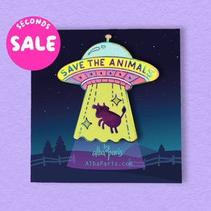 May include: A yellow and pink enamel pin featuring a cartoon UFO abducting a cow. The UFO has the text "Save the Animals" on it. The pin is displayed against a black background with a starry sky and a fence.