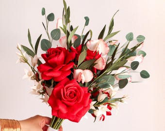 May include: A hand-tied bouquet featuring large red roses, delicate pink and white blooms, and green leaves. The stems are bound with a white ribbon. The arrangement offers a striking contrast of colours and textures.