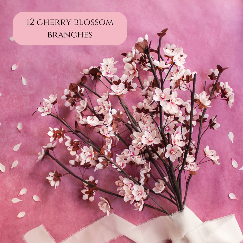 Puede incluir: Doce ramas de cerezo de papel con flores rosas y tallos marrones dispuestas en un ramo sobre un fondo rosa. El texto "12 CHERRY BLOSSOM BRANCHES" est&aacute; en un rect&aacute;ngulo blanco en la parte superior de la imagen.