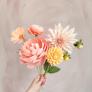 May include: A bouquet of paper flowers in shades of peach, coral, and yellow. The arrangement includes a large peony, a dahlia, and several smaller blooms. The flowers are held in a hand against a soft pink background.