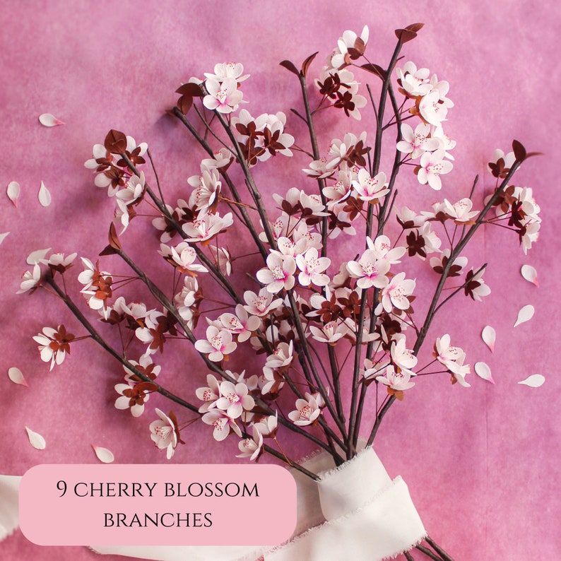 Puede incluir: Nueve delicadas ramas de cerezo en flor con flores rosadas y tallos marrones dispuestas sobre un fondo rosa. Las ramas est&aacute;n atadas con una cinta blanca y el texto "9 RAMAS DE FLOR DE CEREZO" est&aacute; impreso en una etiqueta rosa.