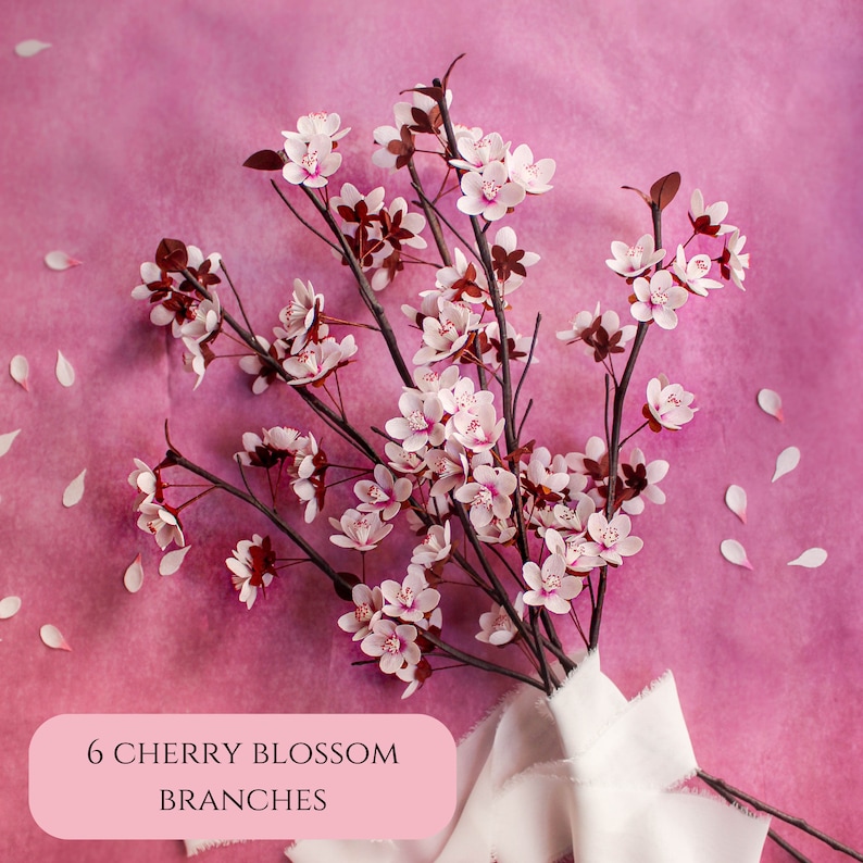 Puede incluir: Seis delicadas ramas de cerezo en flor con flores blancas y tallos marrones dispuestas sobre un fondo rosa. Las ramas est&aacute;n atadas con una cinta blanca. El texto "6 RAMAS DE FLOR DE CEREZO" est&aacute; impreso en un rect&aacute;ngulo rosa.