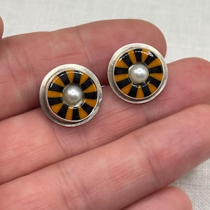 May include: A pair of circular stud earrings featuring a silver-toned metal base. The center of each earring displays a white pearl surrounded by a radial pattern of alternating black and orange enamel stripes, encased within a smooth silver rim.