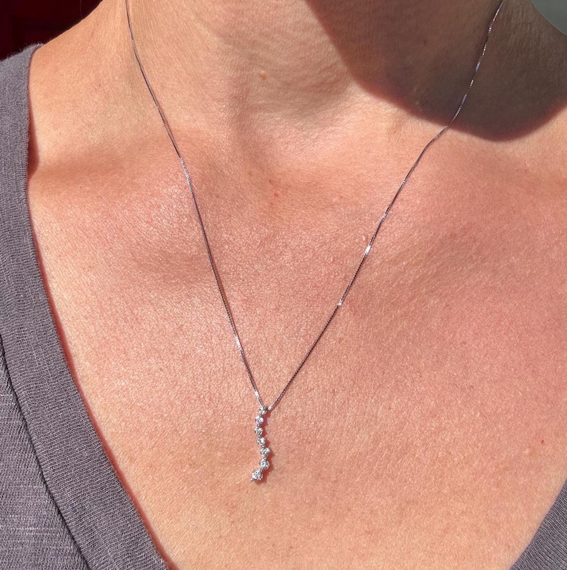 May include: A delicate silver necklace with a series of small, sparkling gemstones arranged in a curved line. The necklace is worn against a gray shirt, creating a subtle contrast. The gemstones appear to be round and evenly spaced.
