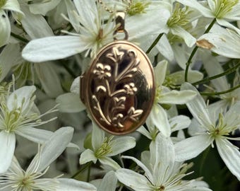 May include: An oval-shaped gold-toned metal locket pendant suspended from a delicate matching chain. The front surface of the locket features an embossed floral design with stems, leaves, and small blossoms centered on the polished metal face.