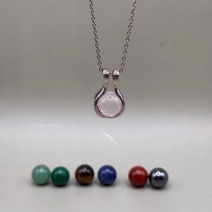May include: A silver necklace with a pendant holding a pink gemstone sphere. Below the necklace are six gemstone spheres in green, dark green, brown, blue, red, and gray.