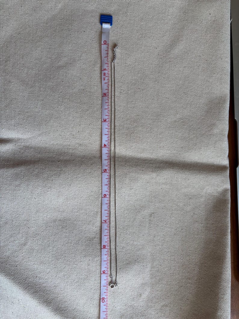 May include: A silver necklace with a small pendant, approximately 10 inches long, is displayed on a beige fabric. A measuring tape is placed next to the necklace, showing its length in inches.