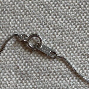 May include: Close-up of a silver necklace clasp and chain. The clasp is a circular ring with a small rectangular tag that reads "10KT" and "KL". The chain is a delicate box chain style.