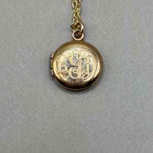 Może przedstawiać: Złoty wisiorek w kształcie medalionu z delikatnym łańcuszkiem. Okrągły medalion ma wygrawerowany monogram z przodu. Medalion jest zamknięty i wisi na szarym tle.