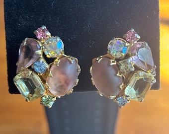Op de afbeelding: Een paar vintage clip-on oorbellen. Elke oorbel heeft een cluster van kleurrijke edelstenen in tinten roze, geel, blauw en iriserend, gezet in een goudkleurig metalen frame. De oorbellen hebben een hartvormig ontwerp.