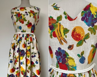 May include: A white linen dress with a fitted bodice and a full skirt. The dress features a colourful fruit print with red, yellow, green, and purple fruits. The dress has a white rope belt and a white tassel.