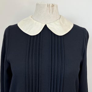 May include: A black long-sleeved dress with a white Peter Pan collar and pleated detail at the front.