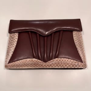 May include: A rectangular clutch bag with a dark burgundy flap and centre panel, featuring vertical pleats. The sides and bottom have a snakeskin pattern in beige and brown. The bag is closed.
