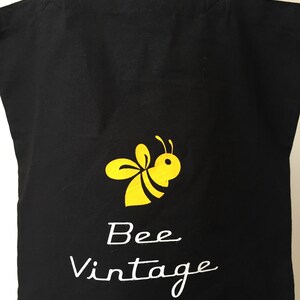 Puede incluir: Bolsa de lona negra con una ilustraci&oacute;n de abeja amarilla y el texto "Bee Vintage" en blanco.