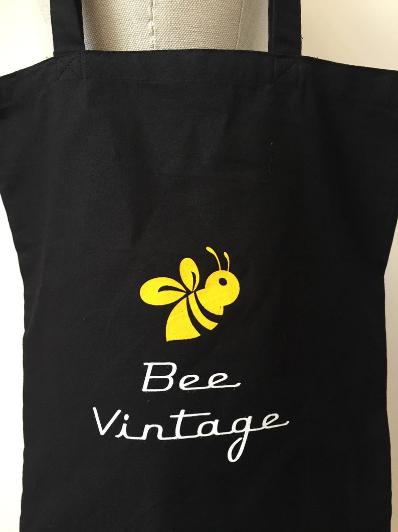 Puede incluir: Bolsa de lona negra con una ilustraci&oacute;n de abeja amarilla y el texto "Bee Vintage" en blanco.