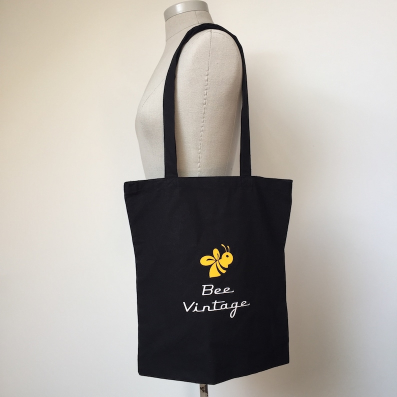 Puede incluir: Bolsa de lona negra con una abeja amarilla y el texto "Bee Vintage" en blanco.
